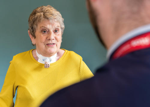 Lady in yellow jumper with a tracheostomy is looking directly at the male nurse in front of her.
