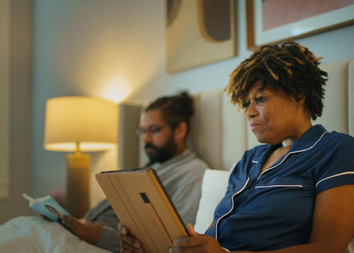 Man and women are sat in bed, man is reading a book and the lady with a laryngectomy is looking at an Ipad.