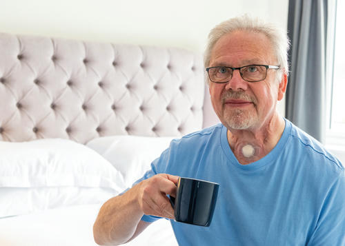 Man with a laryngectomy is sat on his bed wearing a pale blue t-shirt holding a cup of tea.