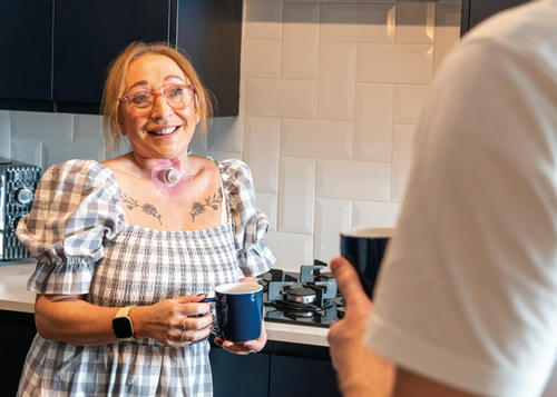 Lady wearing checkered dress with pink glasses is stood in her kitchen holding a cup of tea talking to someone.