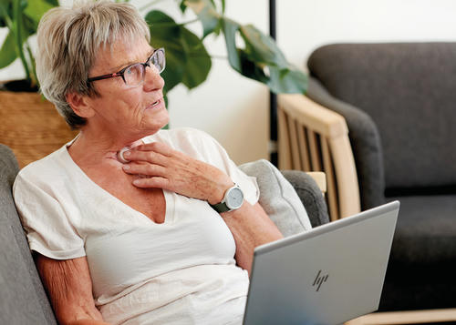 A photo of a lady with a white t-shirt and glasses sat on the sofa with her laptop, pressing down on her HME to occlude voicing.