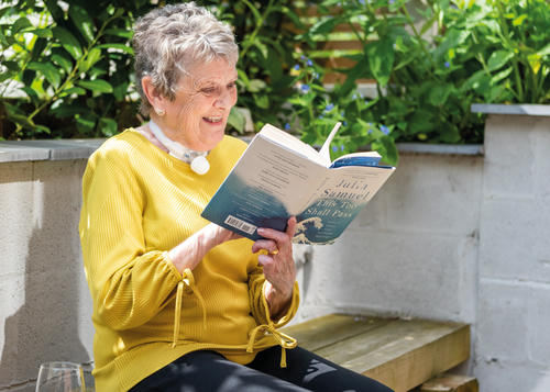 Lady wearing a yellow jumper is sat on a bench in the garden reading her book.