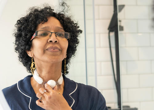 Lady with glasses is looking in the mirror holding her HME on her tracheostomy.